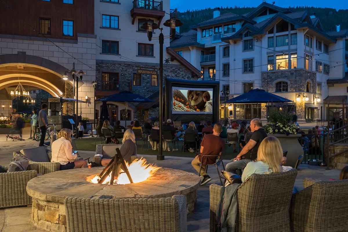 The image captures an outdoor communal space with a fire pit, a projection screen, and a group of guests gathered together. The mood is relaxed and communal, with people enjoying a movie under the stars and sharing stories by the fire, surrounded by a natural setting.