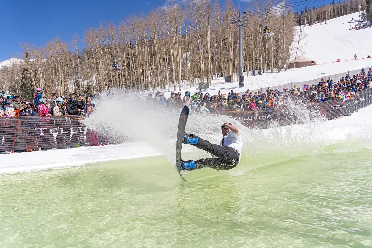 A snowboarder makes a dynamic splash as they land in a pool of water surrounded by snow, with a crowd of spectators and snow-covered mountains in the background. The scene is lively and energetic, capturing a unique outdoor winter event with a festive, community atmosphere.