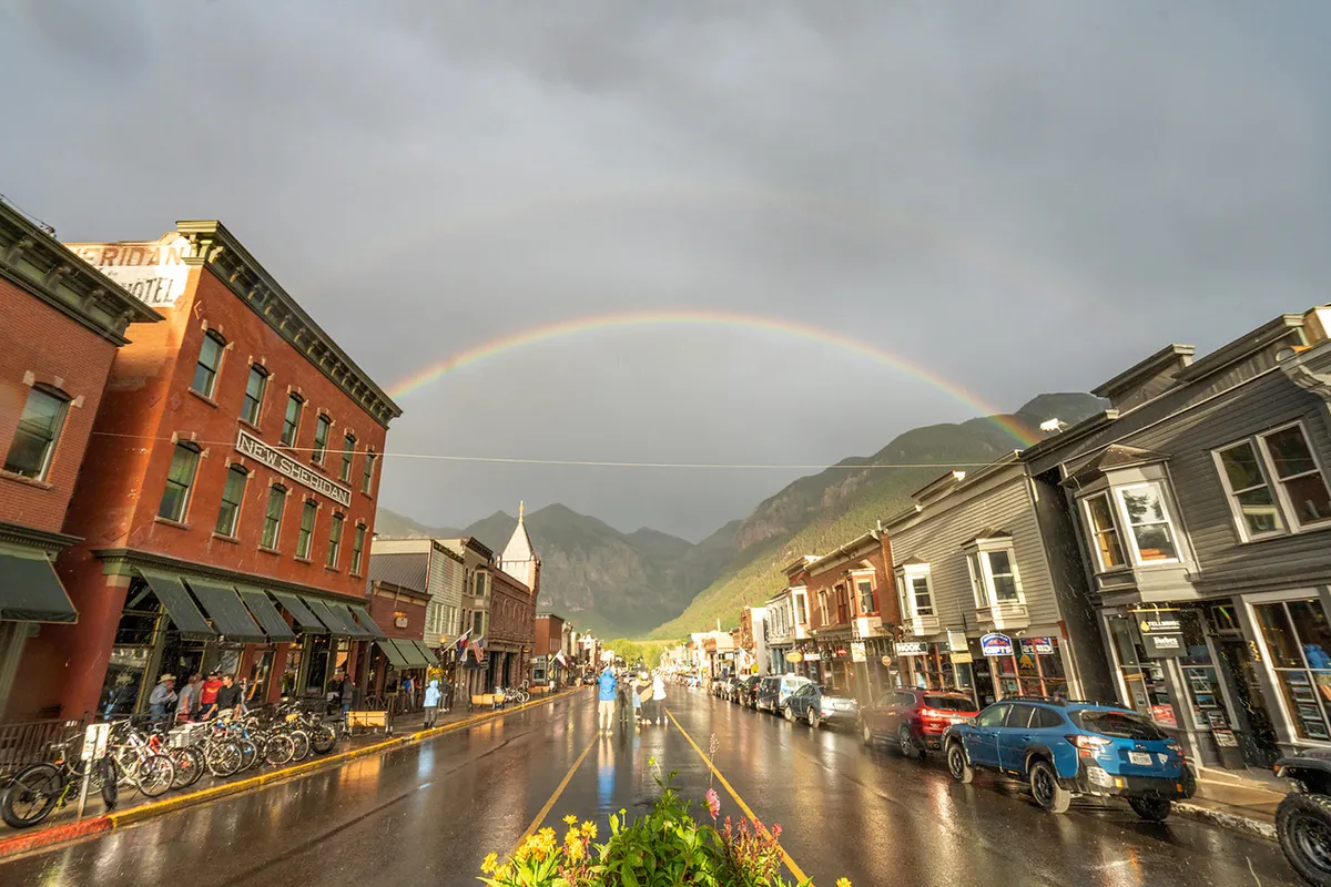 A radiant rainbow arches over historic village buildings against a mountain backdrop, with wet streets reflecting the light and people strolling between unique shops and restaurants.