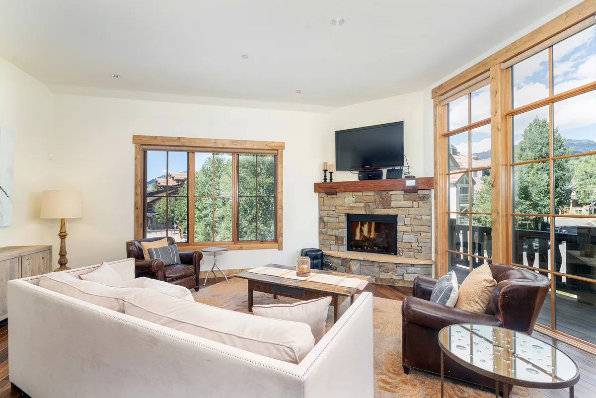 The image showcases a warm and inviting living area with large windows framing scenic mountain views, a cozy fireplace, and plush, comfortable seating arranged for relaxation or gathering with family and friends. Natural light pours in, highlighting the perfect blend of modern comfort and rustic charm.