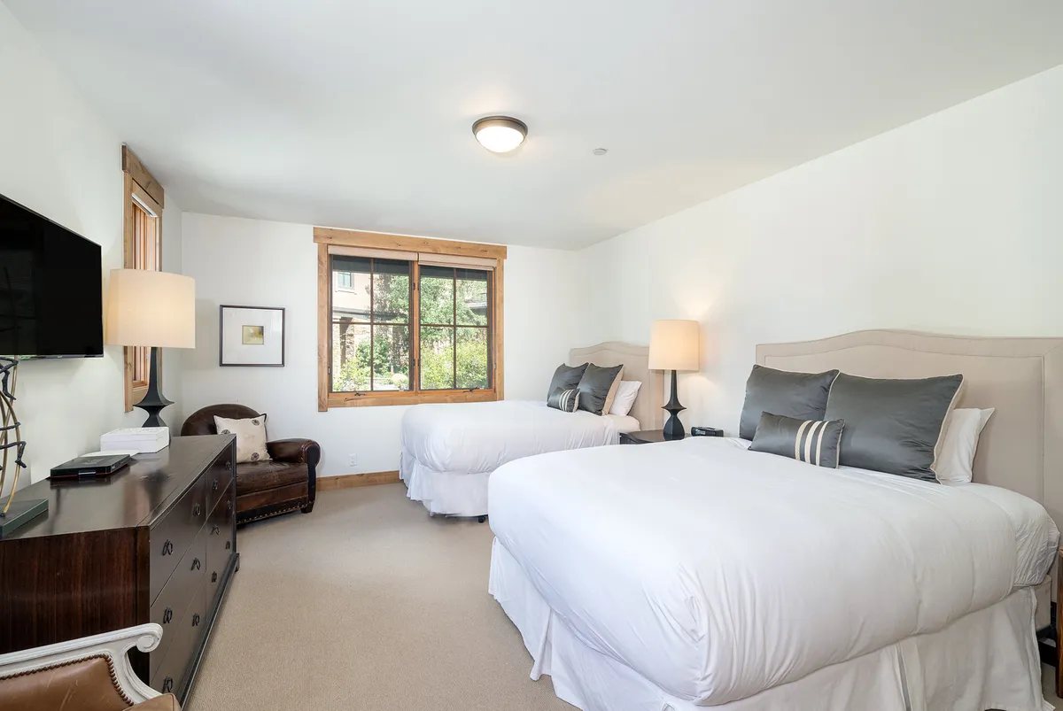A spacious bedroom with two beds dressed in white bedding, wooden window frames letting in natural light, a charming armchair, and a dresser topped with a TV. The overall feel is cozy and comfortable, ideal for relaxing after a day out.