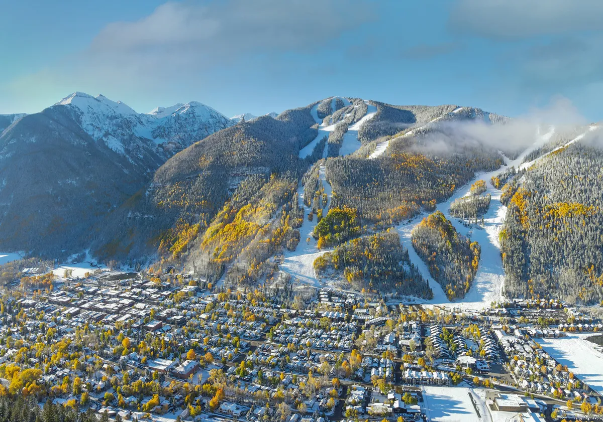 This image showcases snow-capped mountains with ski slopes running down towards a charming mountain town, accented by vibrant autumn trees. The grid layout of the town nestles seamlessly into the landscape, illustrating a close connection between adventure and community life. It gives a broad, panoramic sense of both the natural playground and the nearby après-ski amenities available to guests.