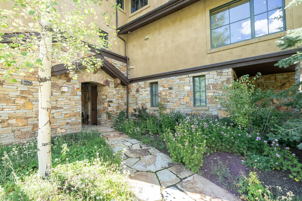 This image shows the stone entrance to the property surrounded by lush greenery, with a clear pathway leading up to the door. Large windows are visible, emphasizing natural light and the welcoming nature of the home. The entrance sets the stage for ease of access and immediate connection to the outdoors.