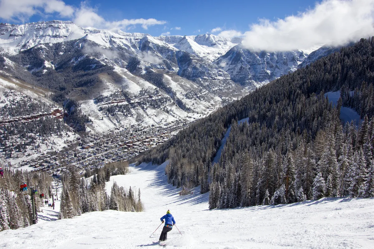 A skier descends a snow-covered slope with a panoramic view of the mountain range and the town below. The scene captures both the thrill of skiing and the closeness of the slopes to the local amenities and accommodations.