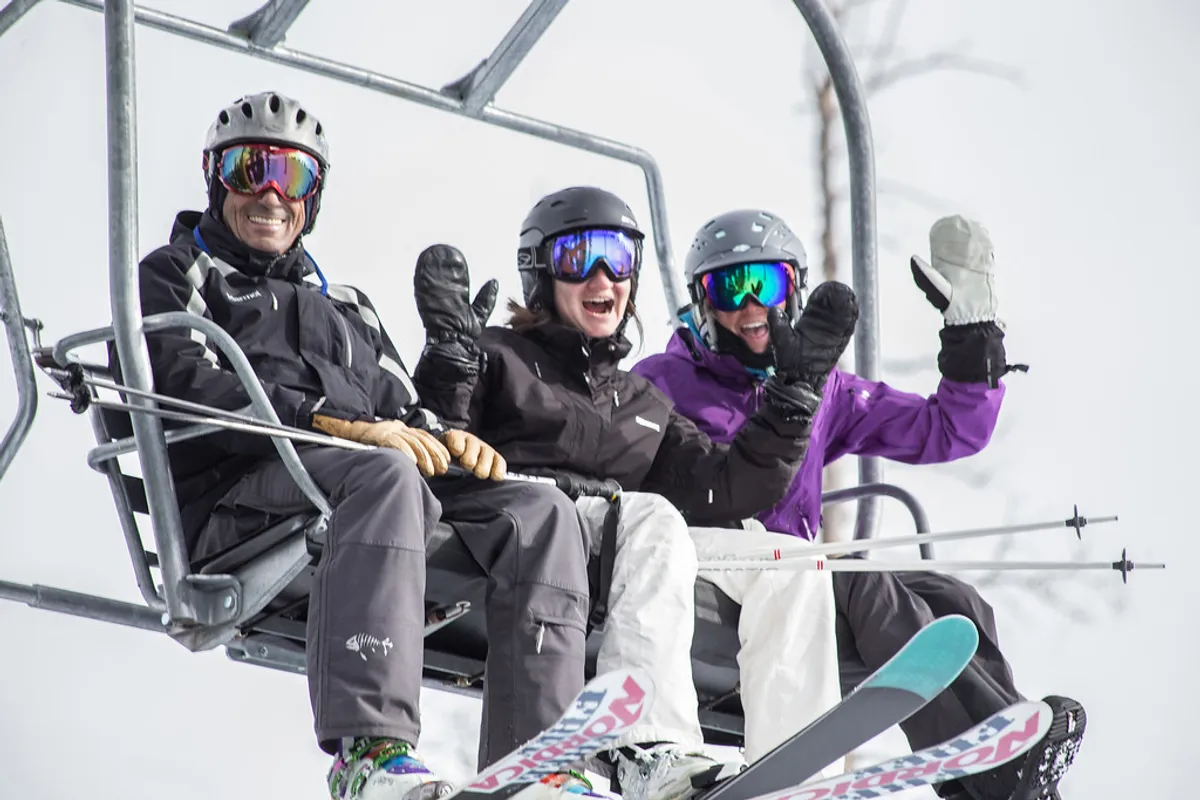 The image features three skiers on a chairlift, surrounded by a snowy mountain landscape. They are fully equipped with ski gear, and the mood is one of excitement and camaraderie, capturing the anticipation of hitting the slopes together.