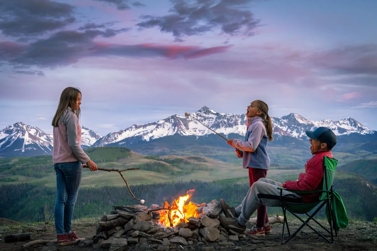 A group gathers around a glowing campfire in front of a majestic mountain backdrop, with children toasting marshmallows and a man relaxing in a chair. The mood is joyful and communal, reflecting evening relaxation and the social possibilities the mountain setting offers in every season.