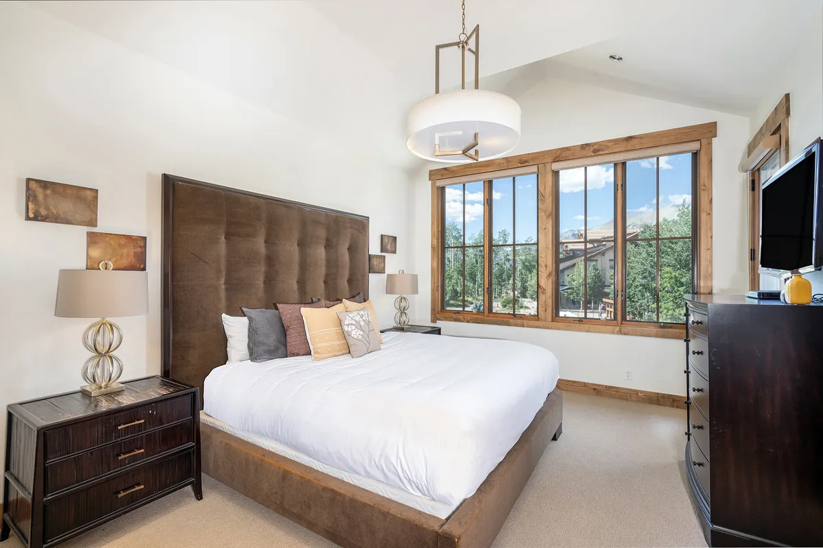 This image presents a spacious bedroom anchored by a large bed with a plush headboard, flanked by nightstands and lamps. Expansive windows allow in natural light and offer views to the outdoors, with decorative pillows and a serene color palette adding to the tranquil environment.