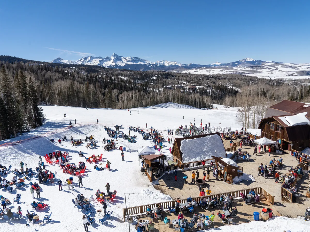 The scene features a lively group of people gathered at the base of snowy slopes, with skiers and snowboarders visible in action. A lodge or hut sits nearby against a dramatic mountain range. The mood is festive and social, suggesting a communal après-ski atmosphere with opportunities for both relaxation and making new friends after a day on the slopes.