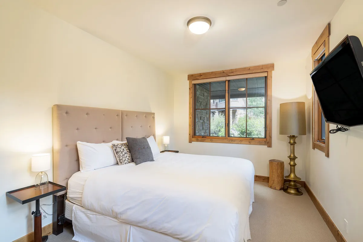 A cozy bedroom scene featuring a comfortable bed placed near a window, a bedside lamp, and a wall-mounted TV, all infused with natural light. The overall vibe is serene and inviting, ideal for unwinding.