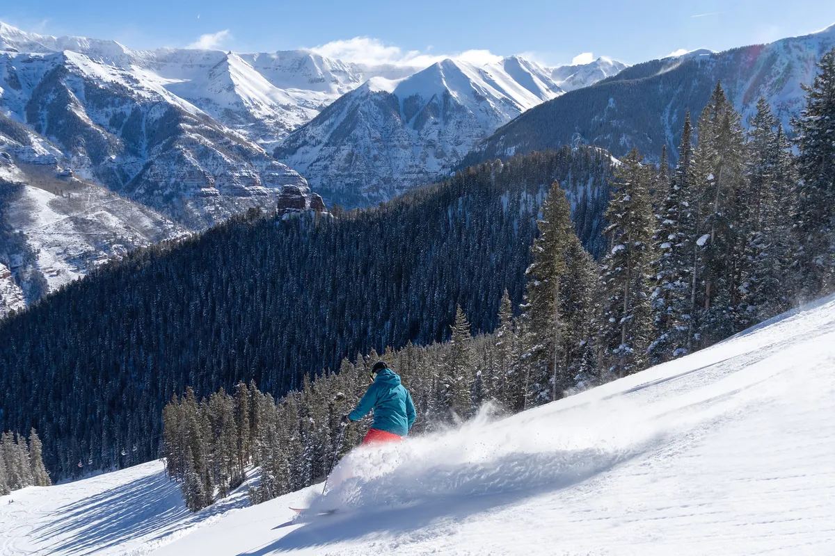 A skier carves down a pristine, snow-covered mountain slope bordered by evergreen trees, with the grandeur of the surrounding peaks forming a dramatic backdrop. The scene radiates energy and adventure, with plenty of open space and natural beauty.