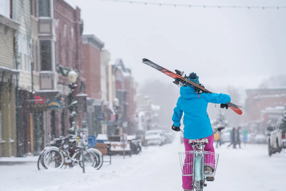 A lively, snow-covered street in the village shows a person biking with skis in tow, historic buildings lining the route, and a playful winter atmosphere. The scene blends the charm of local shops and the unique mountain lifestyle.