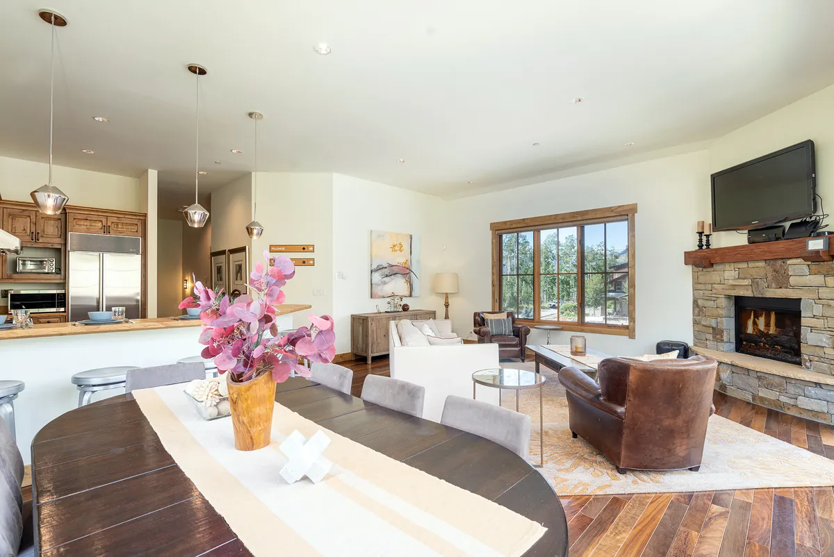This image showcases an open-concept space with a dining table adorned with a floral centerpiece, a cozy seating area with a fireplace, and large windows flooding the room with natural light. The layout encourages gathering, dining, and relaxing, embodying the inviting and communal atmosphere described in the section.